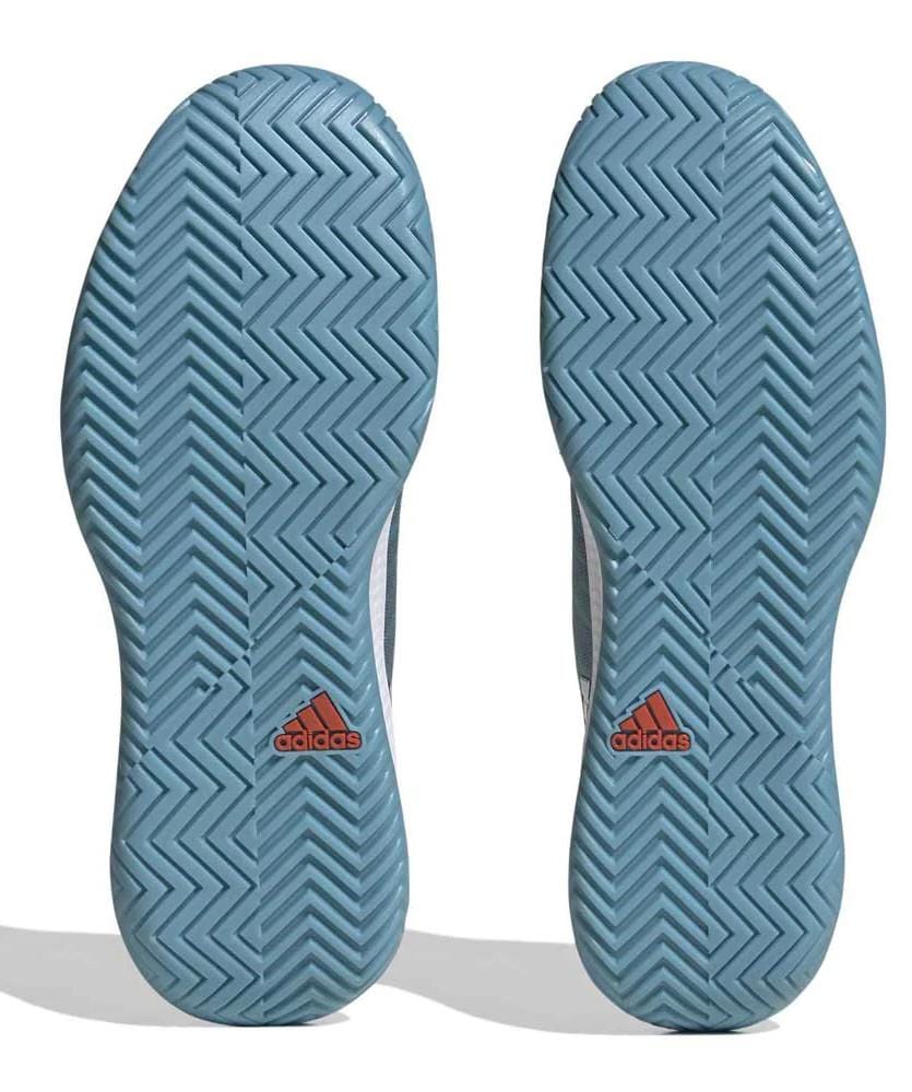 The image highlights the soles of the adidas Defiant Speed women's tennis shoes, prominently featuring a blue chevron tread pattern and a red logo positioned near the heel. Constructed with a lightweight mesh upper, these blue shoes combine style and performance on the court.