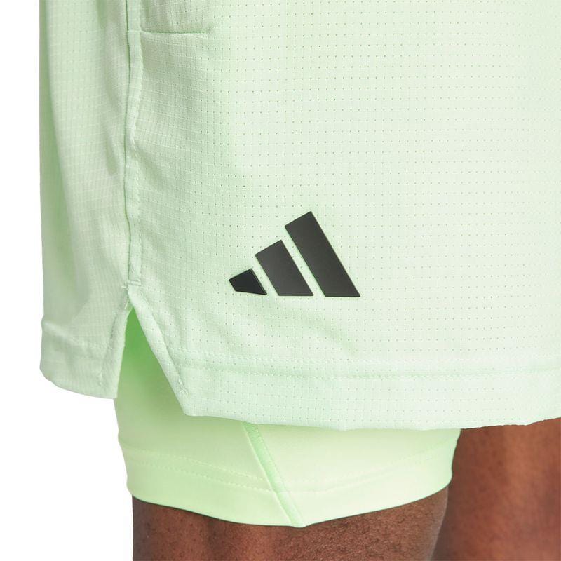 Close-up of a person wearing the light green ADIDAS Melbourne Men's 2in1 Tennis Shorts, featuring a black adidas logo. Made from recycled materials, the shorts have a slightly perforated fabric that reveals a darker green layer underneath and are equipped with HEAT.RDY technology for peak performance.