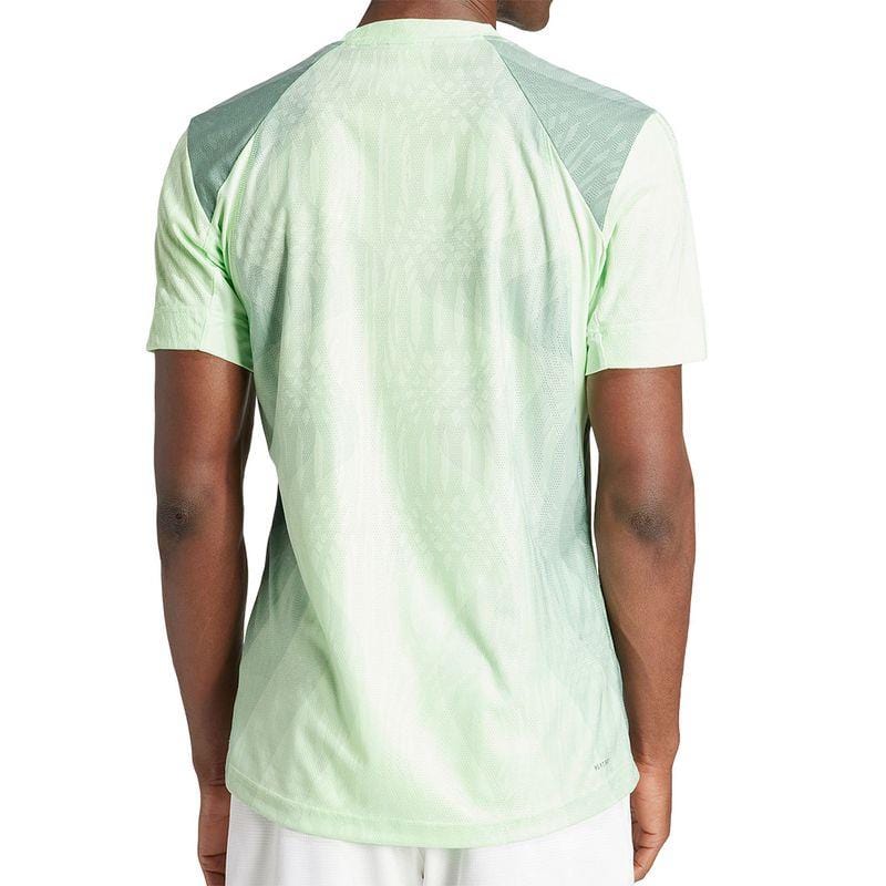 The individual is wearing an adidas Melbourne Men's Pro Tennis T-Shirt in light green with dark green shoulder panels, made from recycled polyester. This HEAT.RDY shirt boasts a subtle pattern and is stylishly matched with white shorts, viewed from the back.
