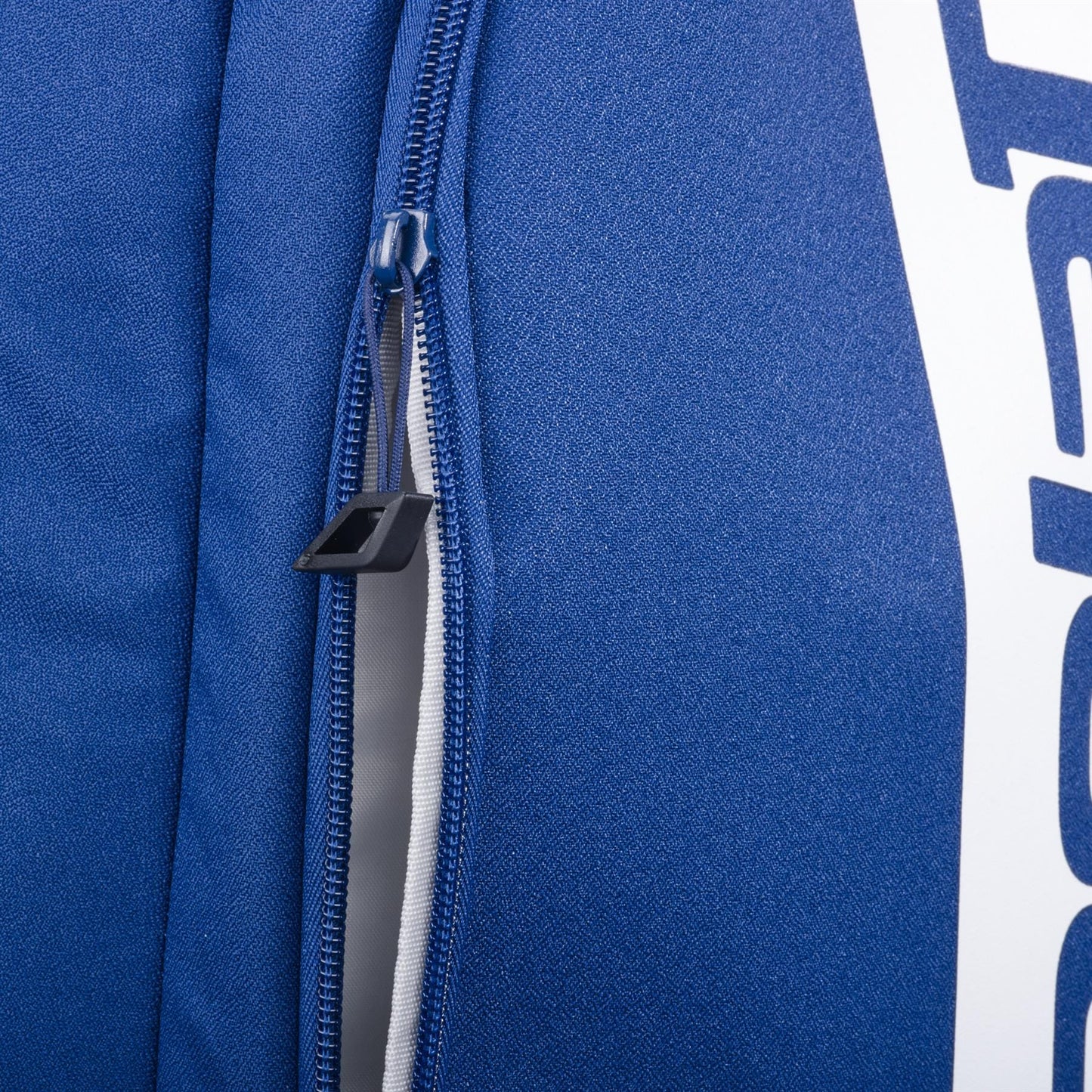 Close-up of the Babolat Court Hero Tennis Backpack - Dark Blue by Babolat, made from recycled PET. The photo shows a partially open zipper with a black pull tab and part of a white printed design on the side.
