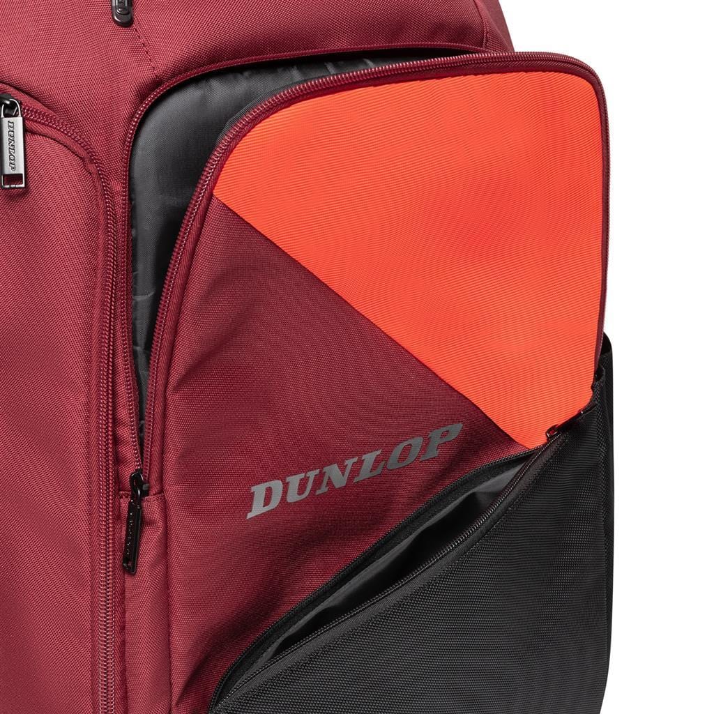 A close-up of the Dunlop CX Performance Tennis Backpack in black and red shows a partially open front pocket with an orange inner lining. The word "Dunlop" is printed in gray on the pocket. This backpack is designed with multiple zippers and compartments, ideal for organizing rackets and gear.