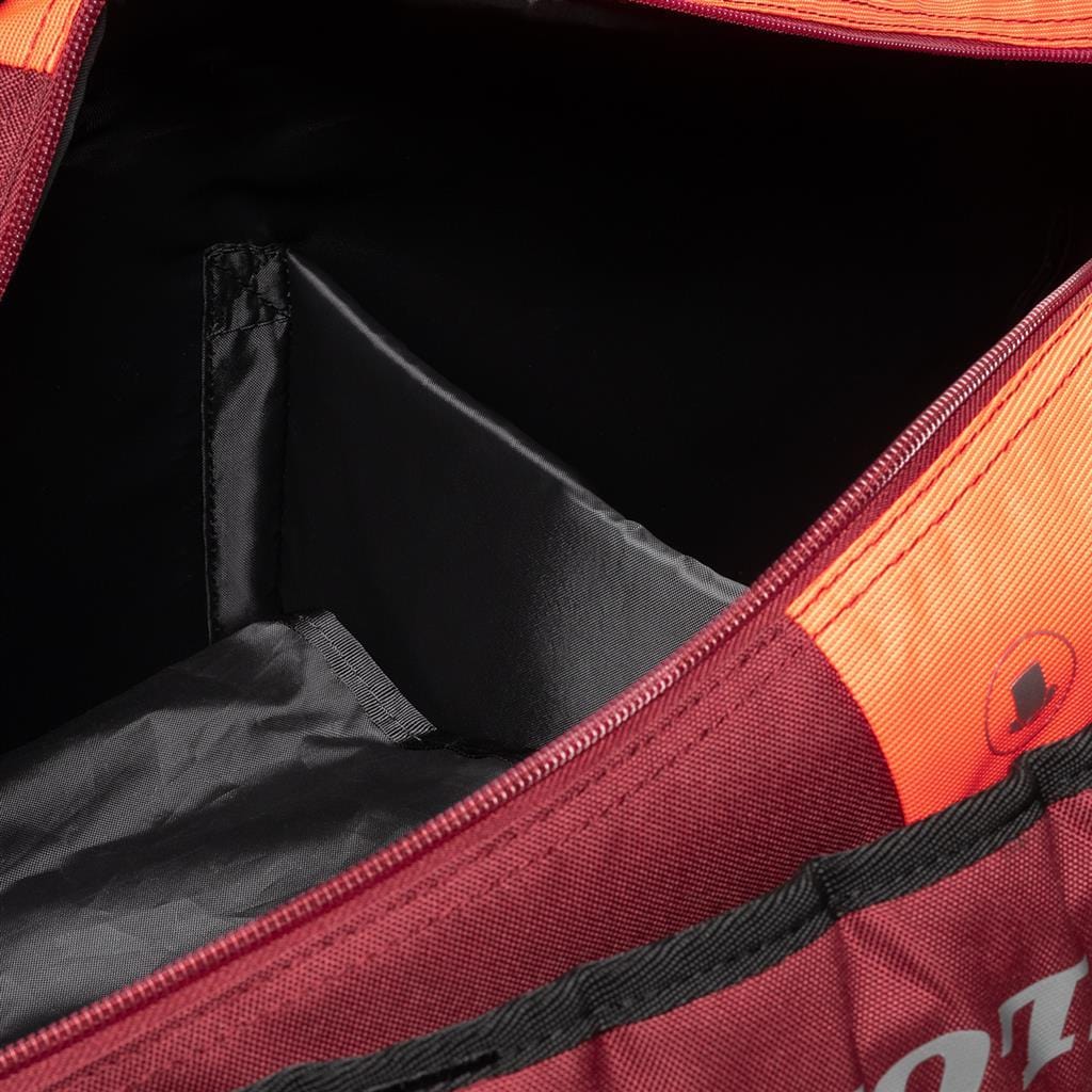 Close-up of an open Dunlop CX Performance 12 Tennis Racket Bag in black and red, displaying its interior compartments. The bag has a partially open zipper that reveals a black lining and a small internal pocket. Designed with ergonomic padded shoulder straps for comfort, it also includes a ventilated shoe compartment.