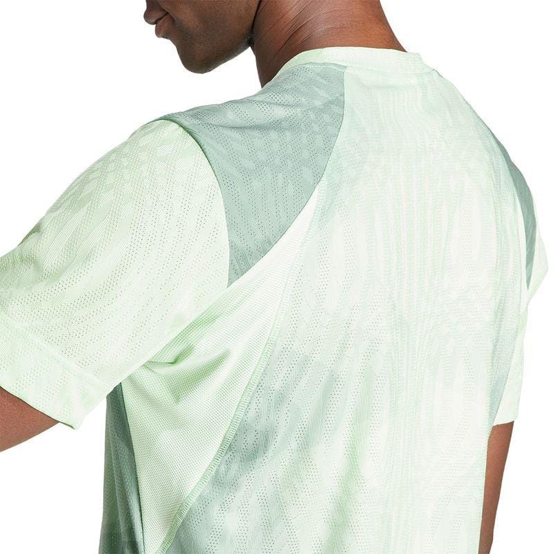 An individual is depicted from behind, their head gently turned, wearing an adidas Melbourne Men's Pro Tennis T-Shirt in green. This eco-friendly shirt is made from recycled polyester and is designed with a mesh pattern on the back and shoulders to incorporate HEAT.RDY technology for enhanced breathability.