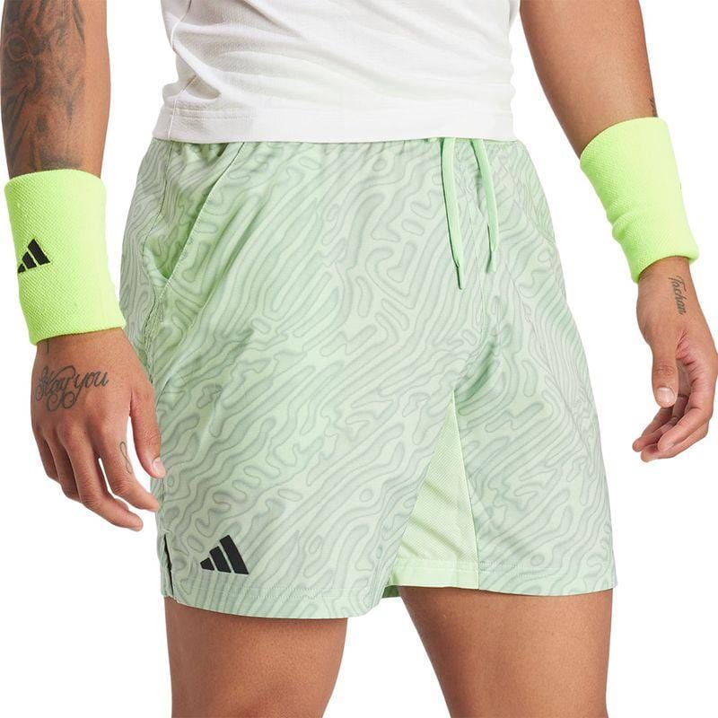 A person is sporting light green ADIDAS Melbourne Men's Pro 7 Inch Tennis Shorts crafted from recycled polyester, paired with a white shirt and neon green wristbands. Both the shorts and wristbands showcase a black adidas sports logo, allowing their tattooed arms to be visible.