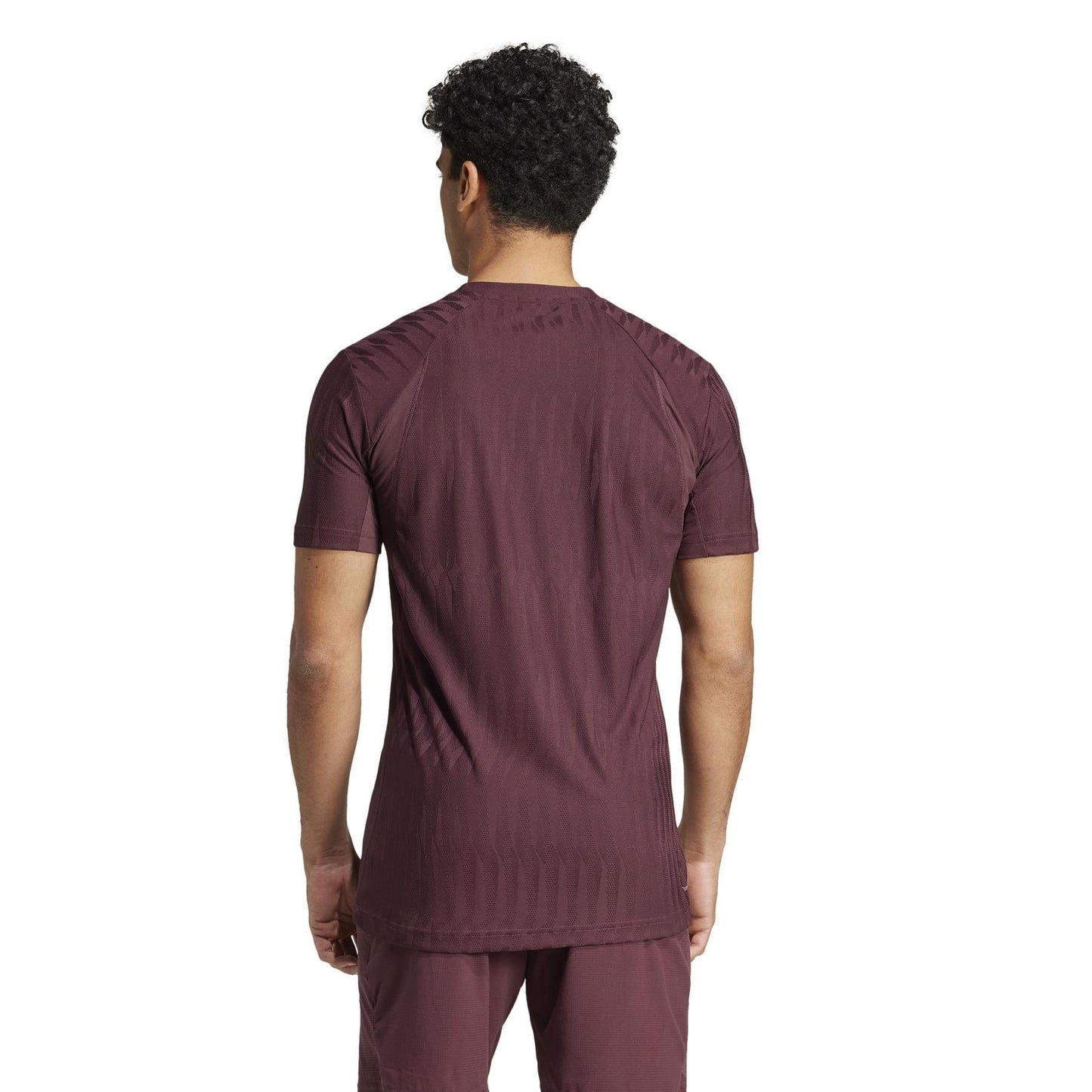 Seen from behind, a person with short curly hair wears an ADIDAS Men's Climacool+ Freelift Tennis T-Shirt in Ruby and matching pants made from recycled materials. The white background emphasizes the sustainable clothing by adidas.