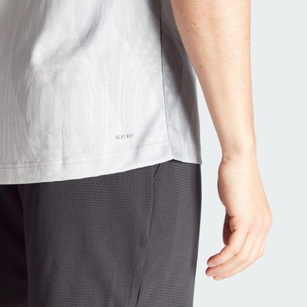A close-up of a person wearing the ADIDAS Melbourn Men's Pro Tennis T-Shirt - Grey by adidas, paired with dark gray shorts, emphasizes the texture and style. Their hand rests naturally, highlighting the shirt's sleek design made from recycled polyester and featuring HEAT.RDY technology.