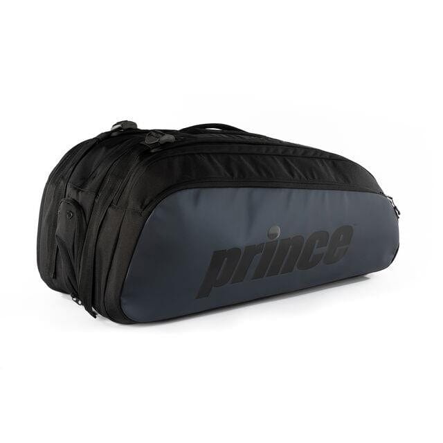 The Prince Tour 3 Comp 12 Racket Tennis Bag - Black, featuring a sleek black design with dark gray accents, boasts multiple compartments including a dedicated shoe compartment and can hold up to 12 rackets. The bag is angled slightly to the left against a plain white background.