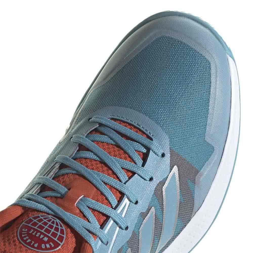 A close-up of the adidas Defiant Speed Women’s Tennis Shoes in blue from adidas showcases its athletic design with a red tongue. This women's tennis shoe features an "End Plastic Waste" logo, highlighting its construction from eco-friendly materials like a lightweight mesh upper. It also includes blue laces and distinct stitching patterns.