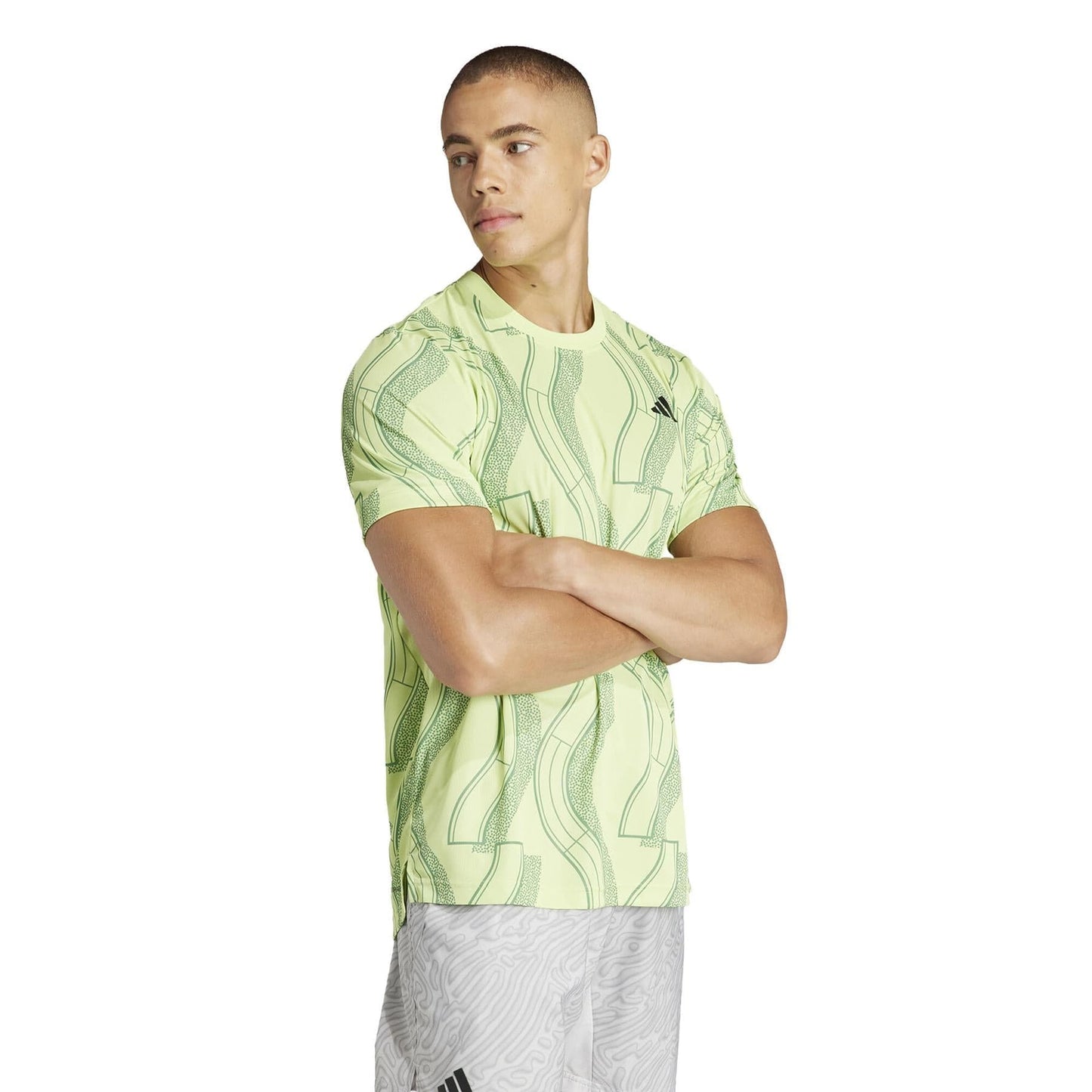 A person stands with arms crossed, wearing a patterned shirt that resembles the ADIDAS Men's Club Graphic Tennis T-Shirt in green and light gray pants. With a neutral expression, they look to the side against a plain white background, showcasing style enhanced by recycled materials.