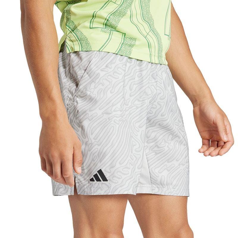 A person wearing a light green patterned shirt and adidas Melbourne Men's Pro 7 Inch Tennis Shorts in grey with abstract designs. The black adidas logo stands out on the shorts, which feature HEAT.RDY technology for cooling comfort and are made from recycled polyester.