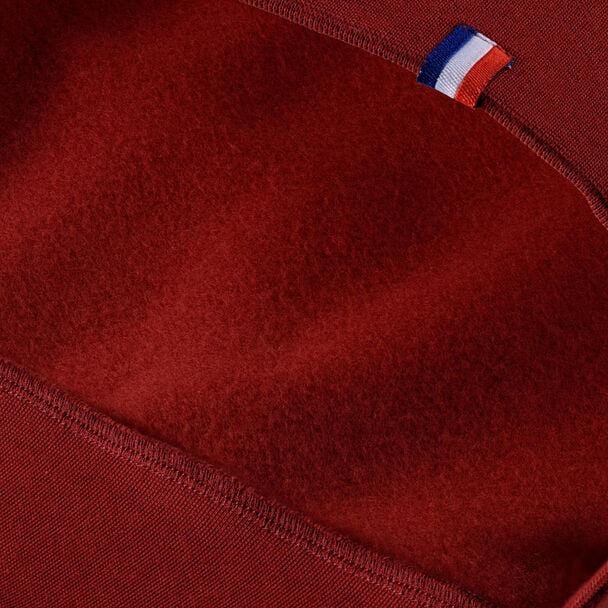 A close-up of a cardinal fabric from the Tecnifibre Men's Team Tennis Sweater reveals two distinct textures. At the top, a small tag featuring blue, white, and red stripes is sewn into the seam of the cotton fleece, adding a detail or branding element that suggests a high-performance style.