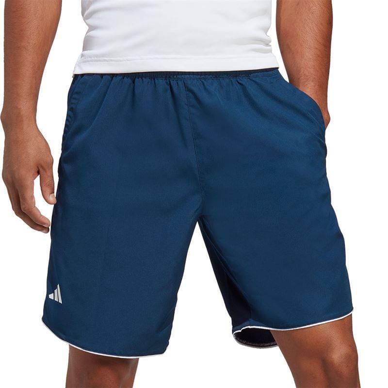 A person wearing a white shirt and dark blue ADIDAS Men's Club 7" Tennis Shorts - Navy is standing. The shorts are made of ripstop fabric and feature a small white logo near the hem.