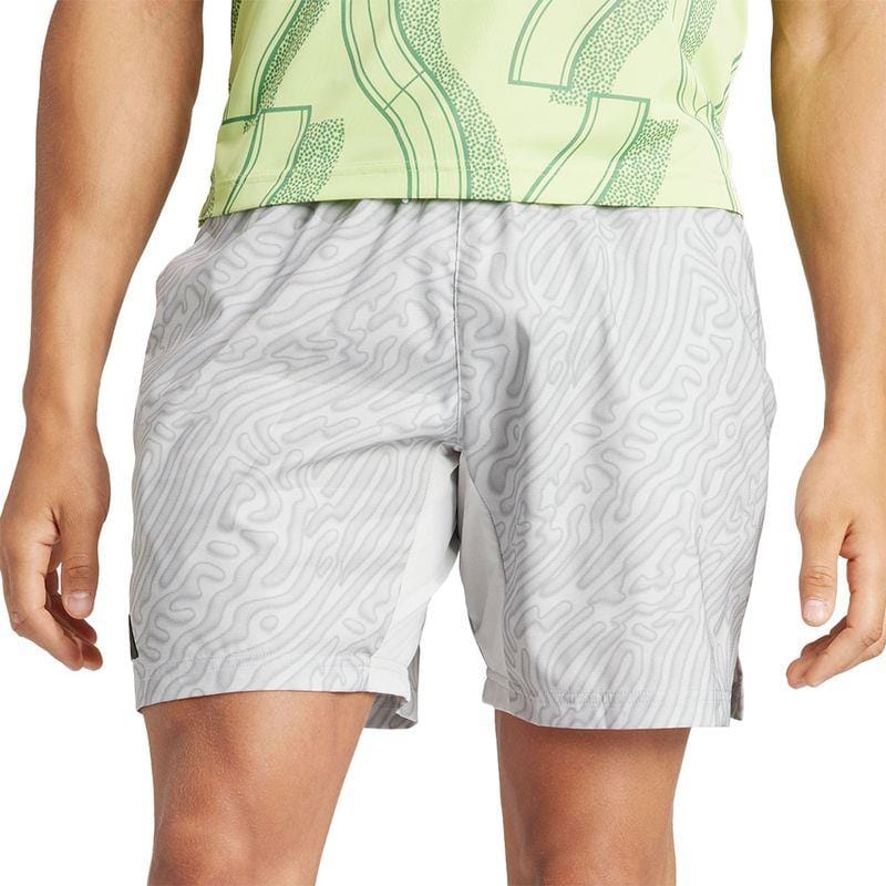 The person is wearing a green patterned shirt along with the adidas Melbourne Men's Pro 7 Inch Tennis Shorts in gray. These shorts, made from recycled polyester, showcase a topographic pattern and HEAT.RDY technology, standing out prominently against the white background.