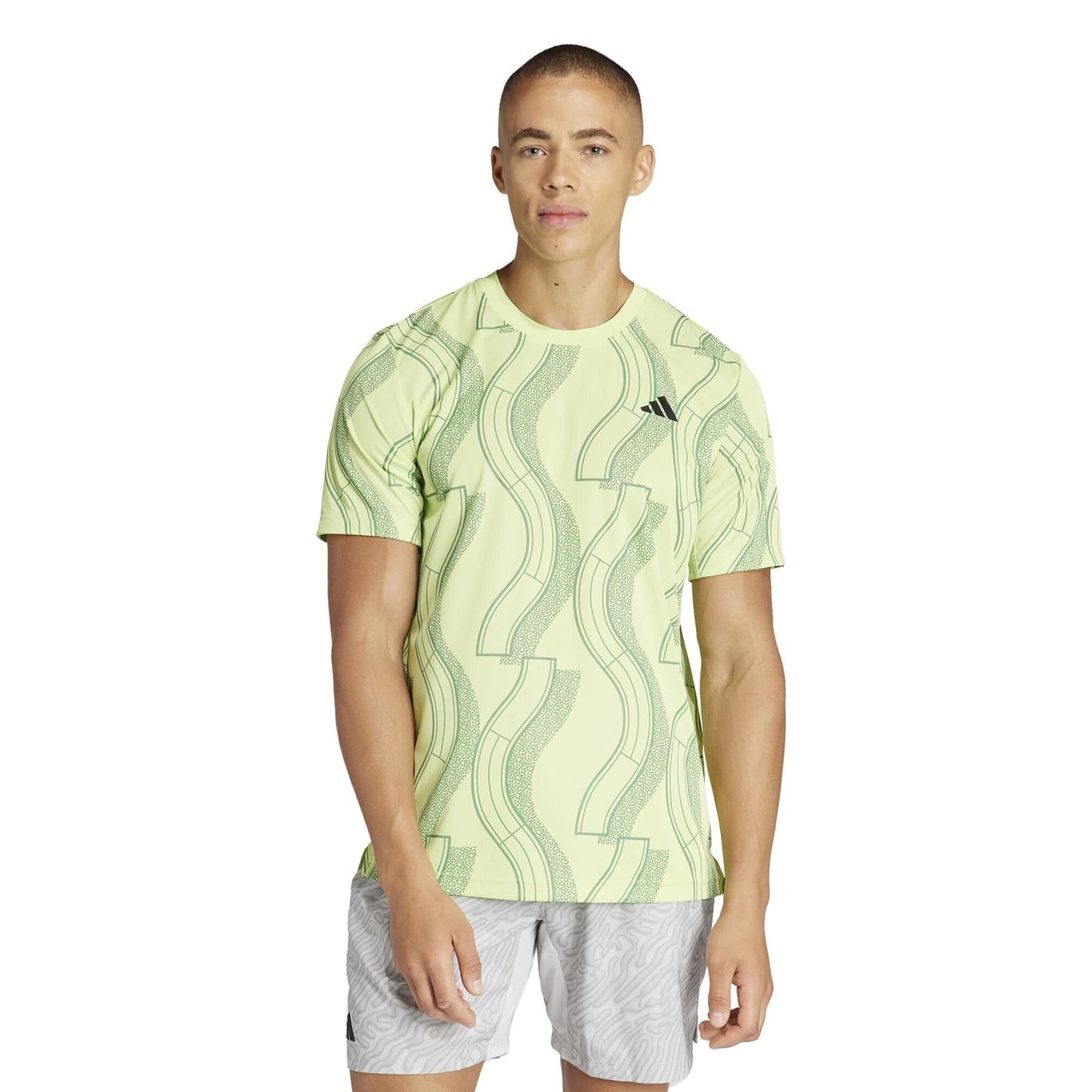 A person wearing an adidas Men's Club Graphic Tennis T-Shirt in green, crafted from recycled materials with abstract green designs, paired with AEROREADY light gray shorts featuring a subtle pattern, stands against a plain white background.