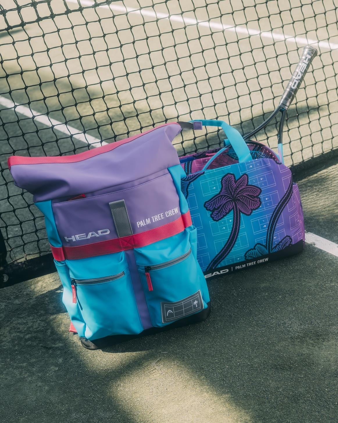 Two colorful tennis bags, including the limited-edition HEAD Tour 30L Tennis Backpack - Palm Tree Crew and a blue and purple duffel with a palm tree design, sit on a tennis court near the net with a racket partly visible in the duffel.