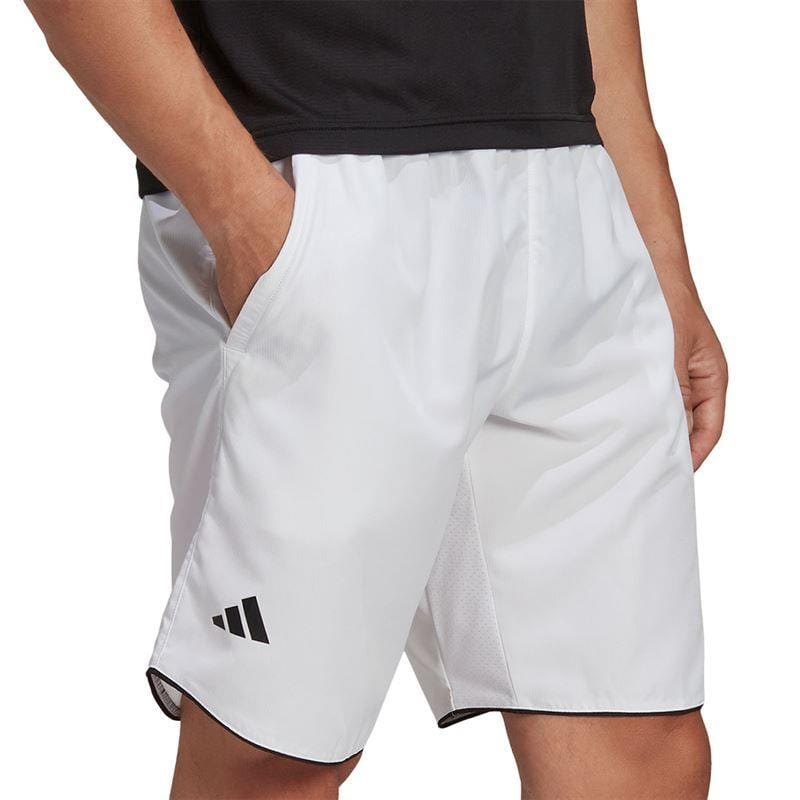 A person wearing ADIDAS Mens Club 7" Tennis Shorts - White with a black logo on the lower left side stands with hands in pockets, paired with a black shirt. These performance-designed shorts by adidas contrast against the plain white background.