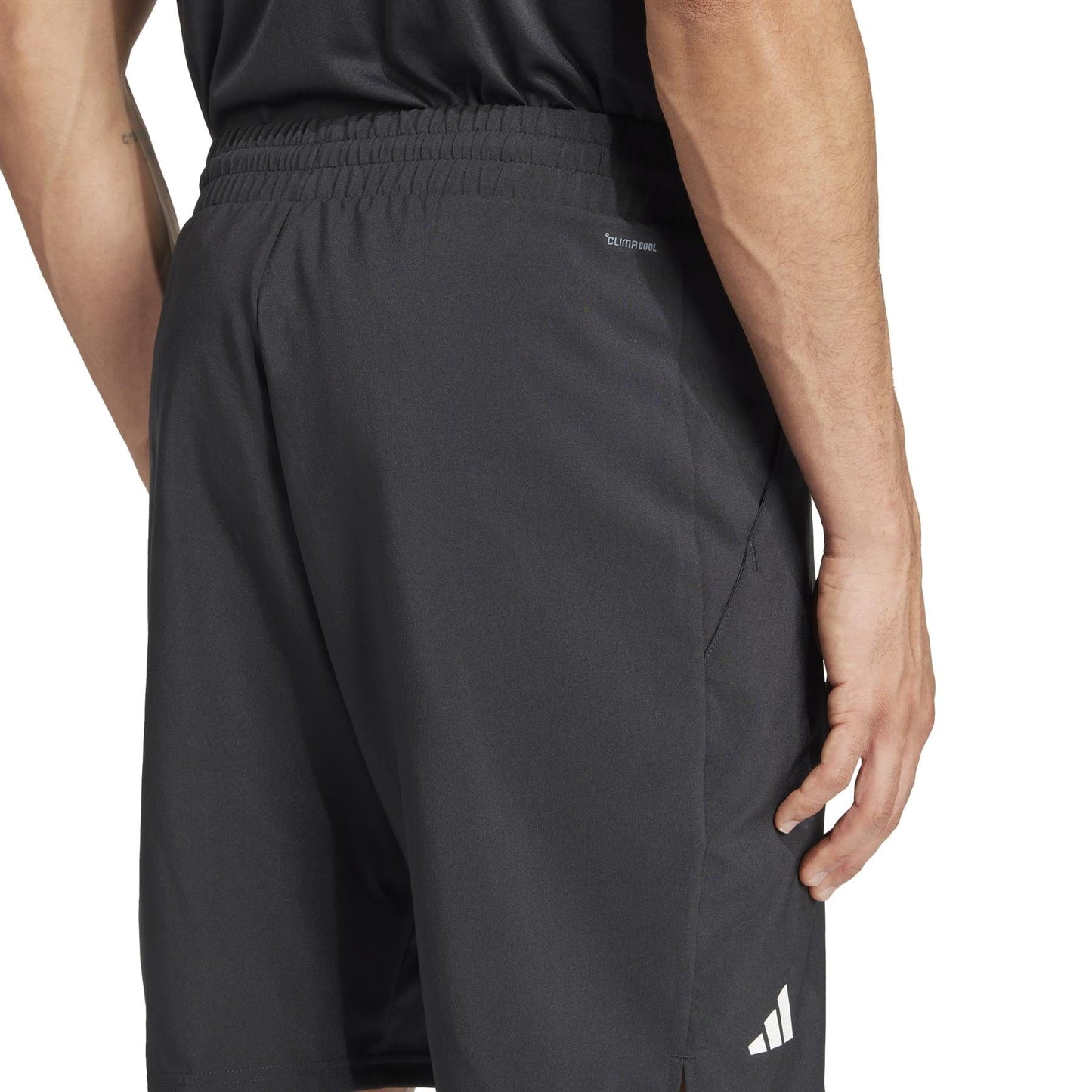 Someone wearing black ADIDAS Men's 3 Stripe Tennis Shorts stands with one visible arm in a plain white background. The shorts have a small white logo near the hem, feature moisture-wicking fabric, and an elastic waistband. They're paired with a partially tucked-in black shirt. .