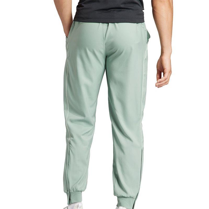 Dressed in ADIDAS Melbourne Mens Pro Tennis Pants in a light green color, and paired with a black shirt for that iconic adidas Melbourne look, the person stands facing away. The pants boast an elastic hem and side stripes designed for AEROREADY comfort. White sneakers finish off this stylish ensemble.