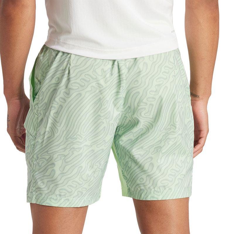 A person in a white shirt and ADIDAS Melbourne Men's Pro 7 Inch Tennis Shorts – Green, designed from recycled polyester featuring a wavy line pattern, stands with their back to the camera. Their right hand rests on their side, prepared for action in HEAT.RDY comfort.