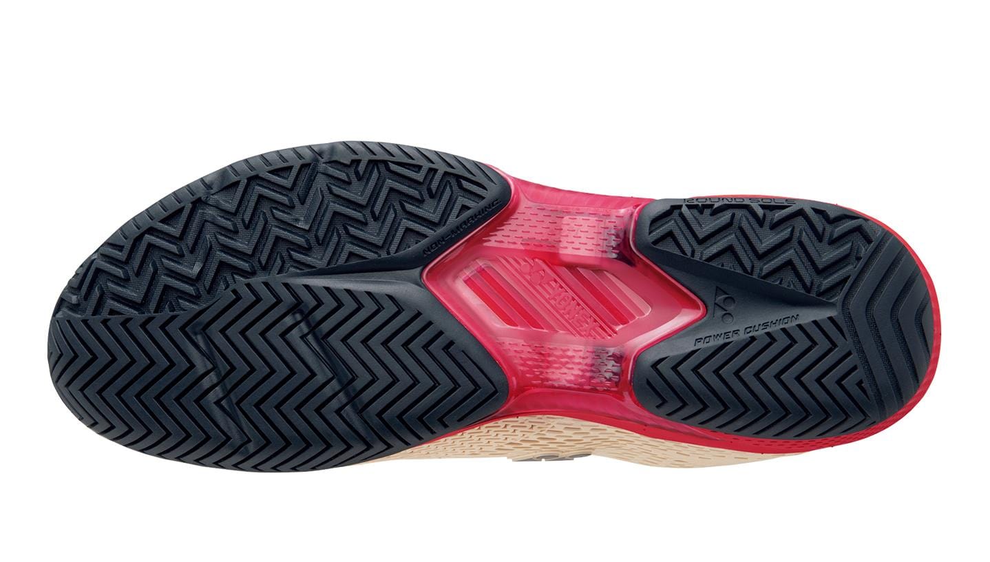 The image showcases the bottom of the Yonex Power Cushion Ad-Accel Men's Tennis Shoes in sand beige, featuring a zigzag tread pattern. The sole is mainly black with red accents and includes Accel-Booster technology and a graphite plate for better cushioning.