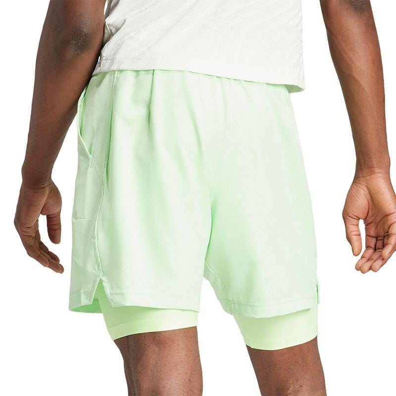 A person wearing the light green ADIDAS Melbourne Men's 2in1 Tennis Shorts from adidas, made with recycled materials, stands facing away from the camera. These shorts showcase layered fabric and a single pocket, incorporating HEAT.RDY technology to ensure maximum comfort. The background is white.