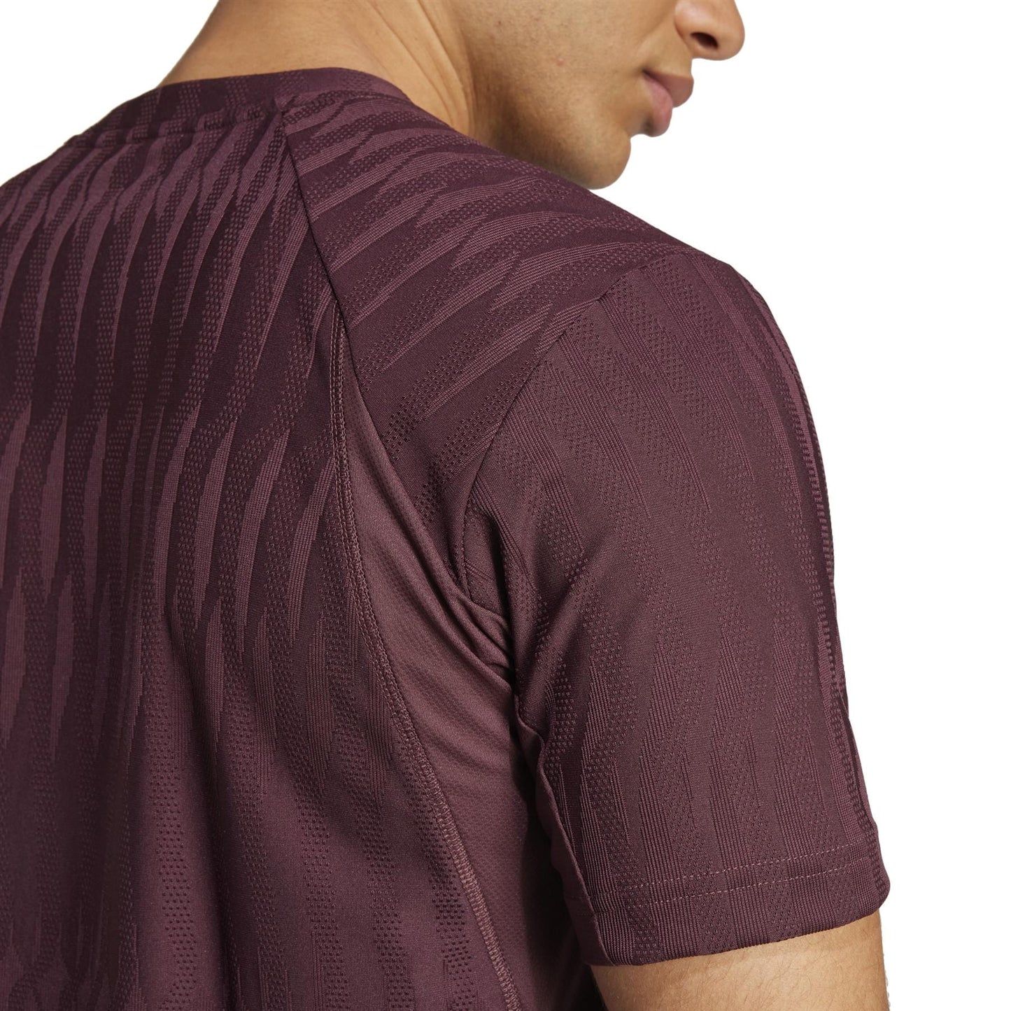 A person is shown from the side wearing an ADIDAS Men's Climacool+ Freelift Tennis T-Shirt in Ruby, made with recycled materials. The focus is on its textured design featuring subtle wave patterns, while the face is mostly out of view.