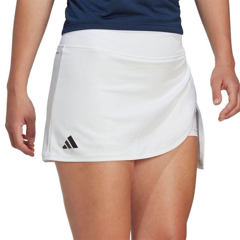 Dressed in a blue top and an ADIDAS Women's Club Tennis Skirt - White with a wide waistband, subtly adorned with a small black logo on the side, an individual stands against a plain background. The skirt features a wrap design crafted from AEROREADY fabric, complete with a slight slit on one side.