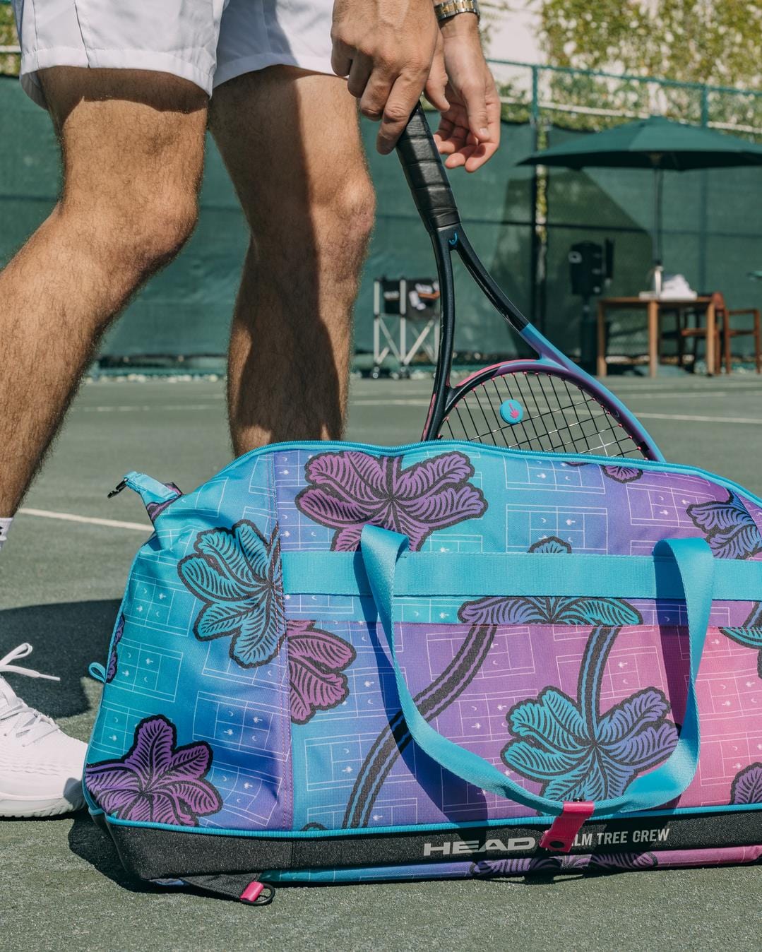 A person in white tennis shorts and sneakers stands on a court with a HEAD Tour Tennis Duffle Bag L - Palm Tree Crew by HEAD, carrying a tennis racket. In the background are a net, chairs, and outdoor equipment.