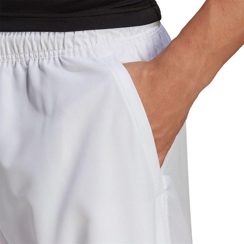 A close-up shows a person wearing the ADIDAS Men's Club 7" Tennis Shorts in white, crafted from ripstop fabric, with their right hand tucked into the pocket. They are also sporting a black top. The emphasis is on the shorts and pocket detail, showcasing AEROREADY technology for enhanced comfort.