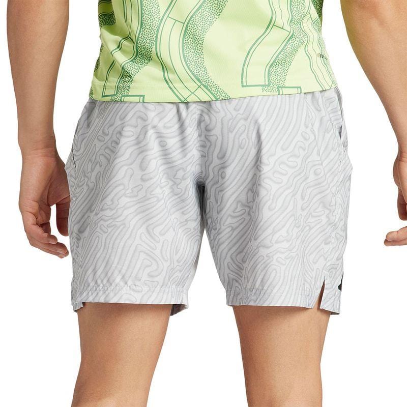 A person, wearing the ADIDAS Melbourne Mens Pro 7 Inch Tennis Shorts - Grey crafted from recycled polyester, stands facing away from the camera. The light gray shorts showcase a wavy design, complemented by a light green patterned top optimized with HEAT.RDY technology for maximum comfort.