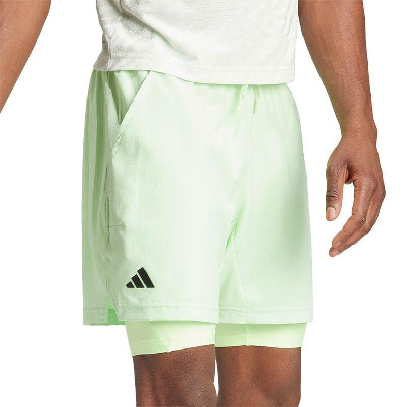 A person is wearing the ADIDAS Melbourne Mens 2in1 Tennis Shorts in a light green color by adidas. These shorts are made from recycled materials and feature an overlapping inner layer, showcasing a black logo on the left leg. They pair perfectly with a matching light green t-shirt, emphasizing their HEAT.RDY technology for peak performance.
