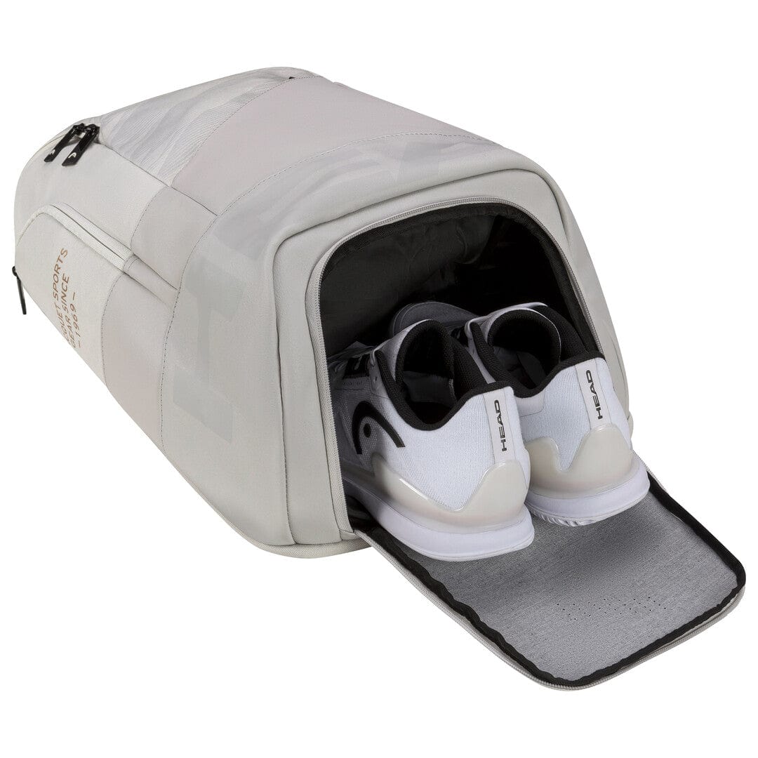 A HEAD Pro X 28L Tennis Backpack in gray features a ventilated shoe compartment revealing a pair of white sneakers. The backpack is equipped with a zipper and showcases visible stitching details, perfect for those who value organization and style.