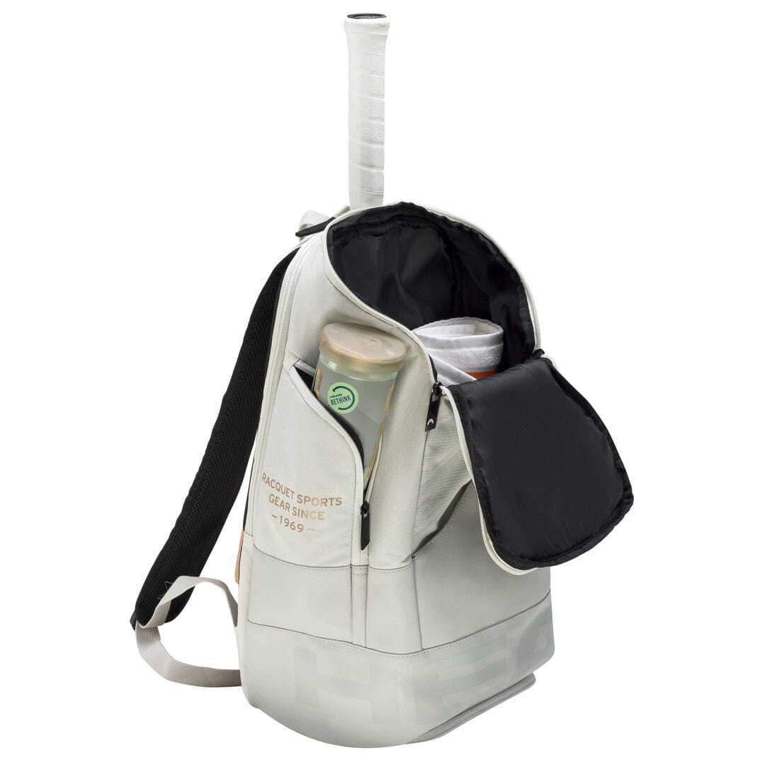 The HEAD Pro X 28L Tennis Backpack - YUBK, in a white design, showcases a racquet handle protruding from the top and features an open front pocket exposing tennis balls alongside a canister. Equipped with a padded racquet compartment for added protection, the bag also includes black straps adorned with the phrase "Racquet Sports Gear Since 1969.