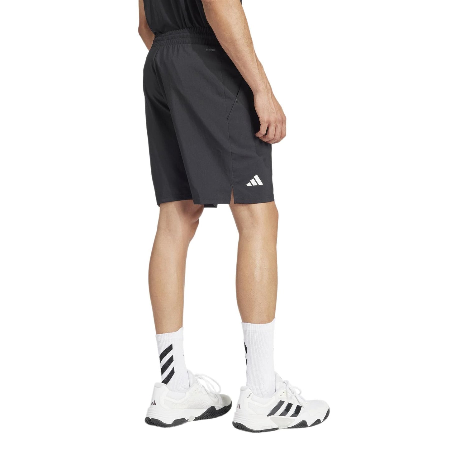 A person is wearing black ADIDAS Mens 3 Stripe Tennis Shorts - Black, a black top, and white sneakers with long white socks, standing against a white background. The shorts showcase the adidas logo on the left and are made from moisture-wicking fabric for comfort.