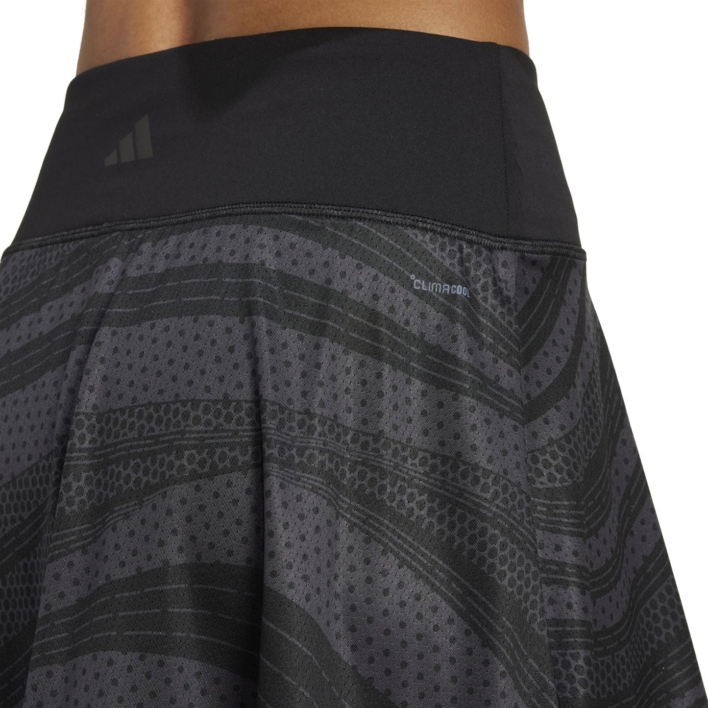 Close-up of a person wearing the ADIDAS Womens Club Graphic Tennis Skirt - Black, featuring a dark-patterned design with a black waistband. The recycled fabric boasts subtle stripes and dots, with the "climacool" label and emblem visible on the waistband.