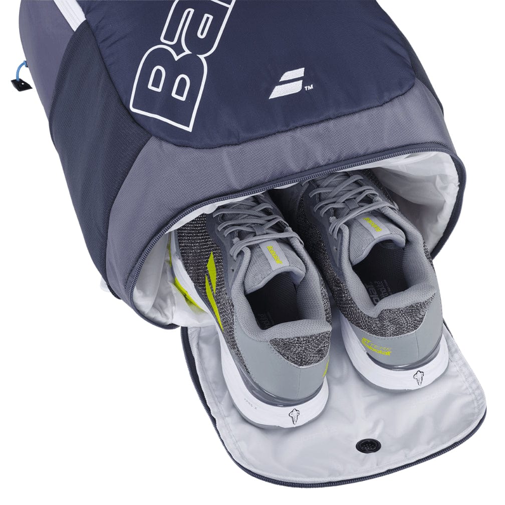 A pair of gray and white athletic shoes with neon yellow accents tucked inside the shoe compartment of a Babolat Evo Court Tennis Backpack in grey and blue, crafted from eco-friendly materials.