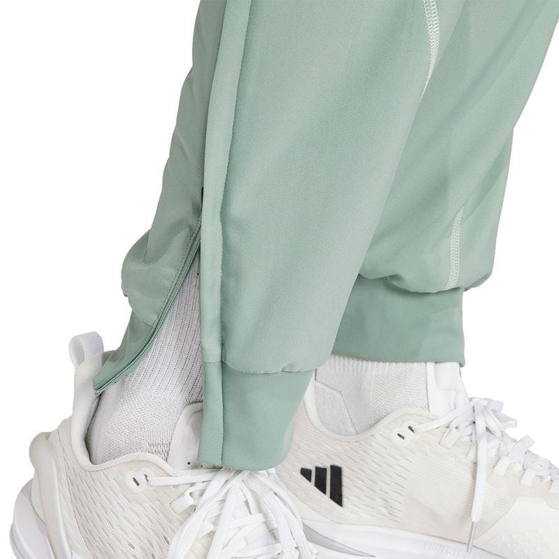 A close-up shot shows an individual donning the ADIDAS Melbourne Men's Pro Tennis Pants in a light green shade, paired with white sneakers. The pants, designed by adidas, feature zippered ankles and a sleek design. The sneakers are accented with black striped details. AEROREADY technology is prominently showcased in the lower leg area of the pants and on the shoes.