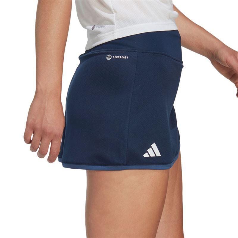 The image shows an individual wearing a white shirt paired with the ADIDAS Women's Club Tennis Skirt in navy, featuring a discreet logo on the side. The focus is on the lower body from a side view, highlighting the skirt's sleek athletic design and AEROREADY fabric.