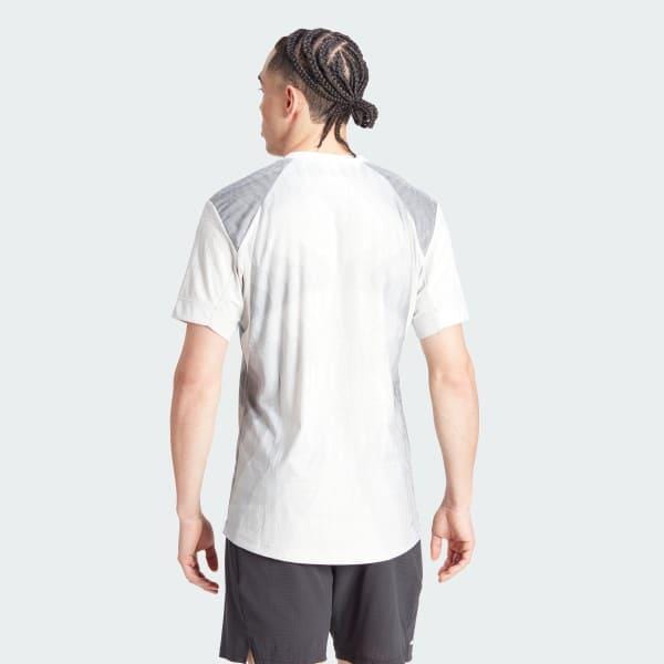 Against a plain backdrop, an individual with braided hair is shown from behind, wearing the adidas Melbourne Men's Pro Tennis T-Shirt in grey, crafted from recycled polyester. This light-toned sports tee showcases HEAT.RDY technology and darker shoulder panels, complemented by dark shorts.