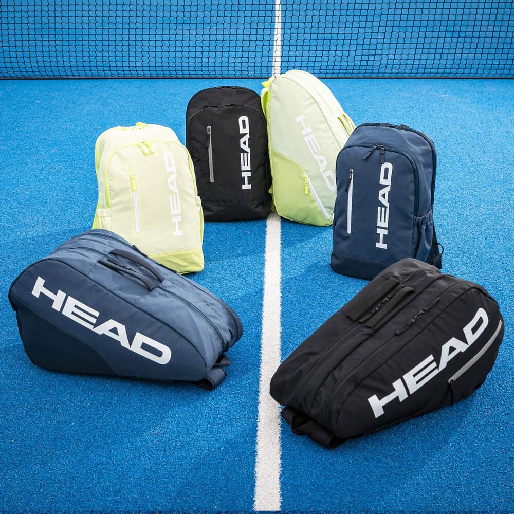 Six HEAD tennis bags, including the HEAD BASE Tennis Backpack 17L - Navy with a new design and racket compartment, are arranged in a semicircle on the blue tennis court near the net—three upright and three lying down.
