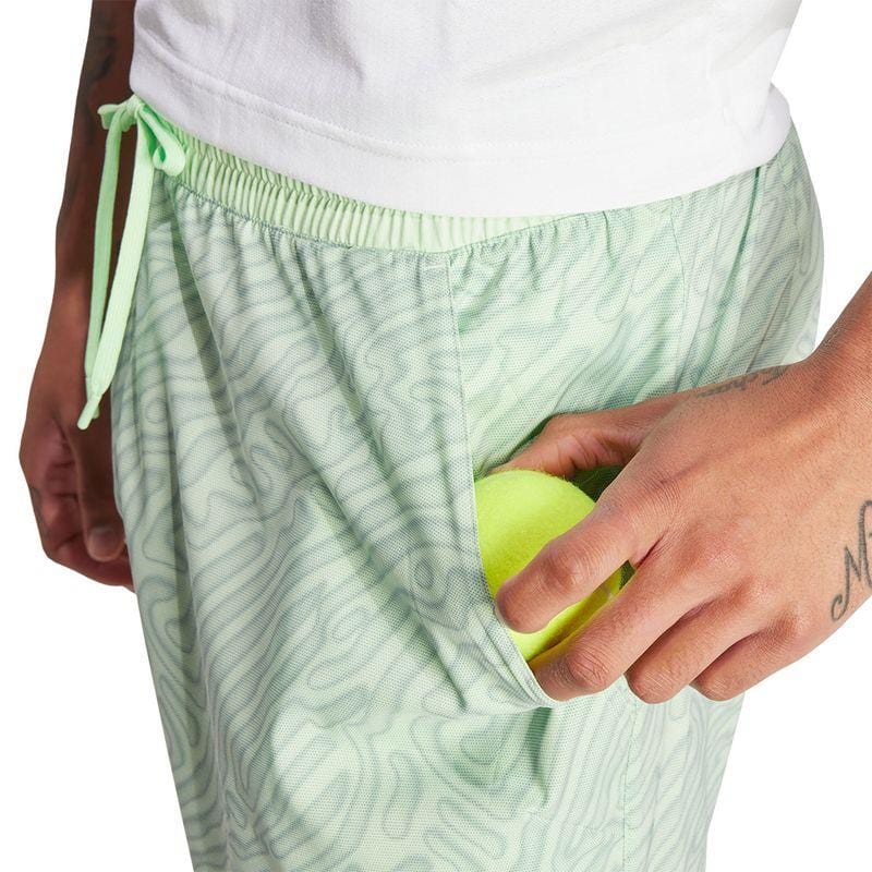 Dressed in the ADIDAS Melbourne Men's Pro 7 Inch Tennis Shorts in a vibrant green shade, designed from recycled polyester for eco-friendliness and featuring HEAT.RDY technology for optimal cooling, a person is tucking a tennis ball into their right pocket. The shorts are accentuated by a light green drawstring and paired with a white shirt. Their forearm and hand showcase intricate tattoos.