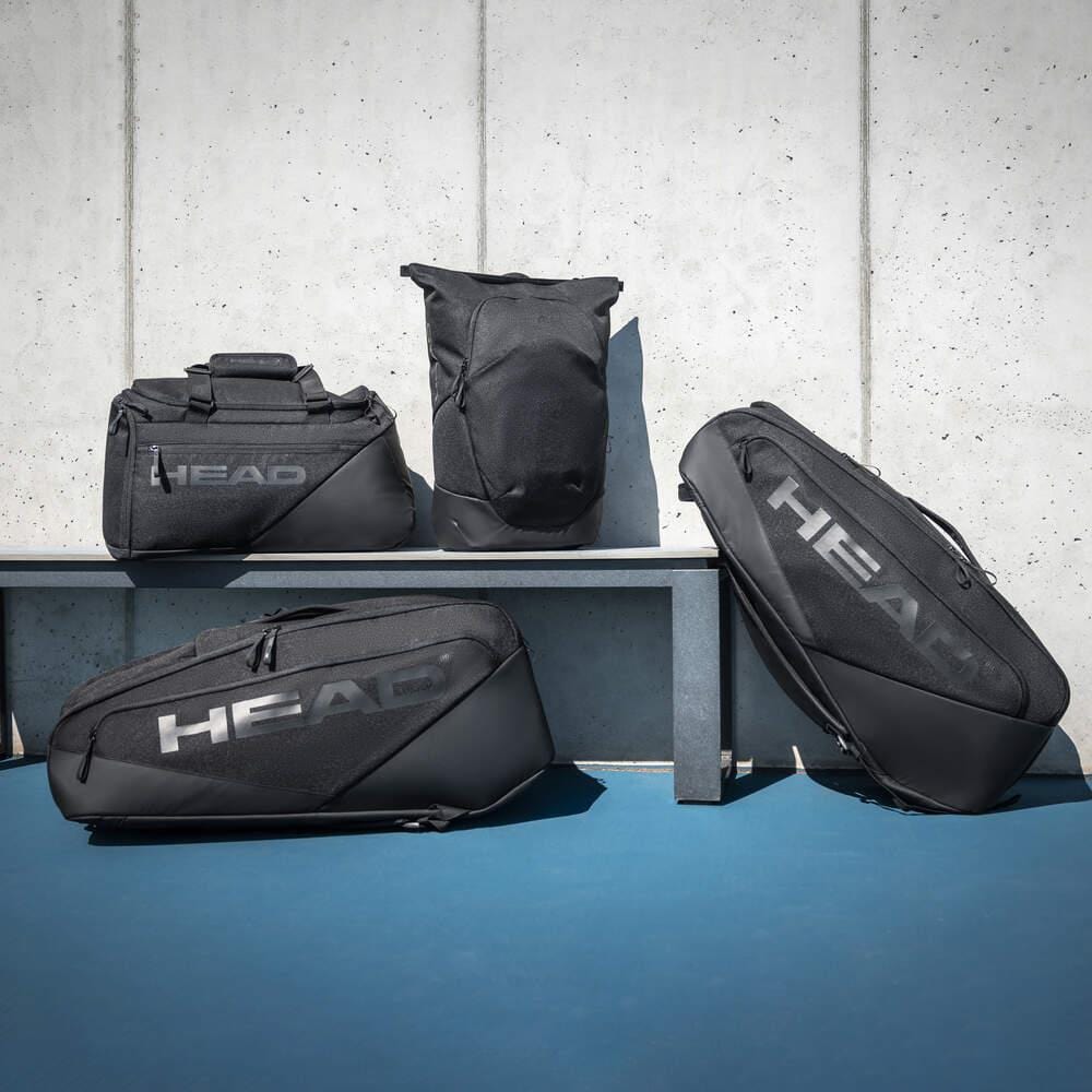 Four black HEAD Pro X 12 Racket Tennis Racket Bag XL by HEAD are displayed on and in front of a gray bench against a concrete wall, with a blue floor below.