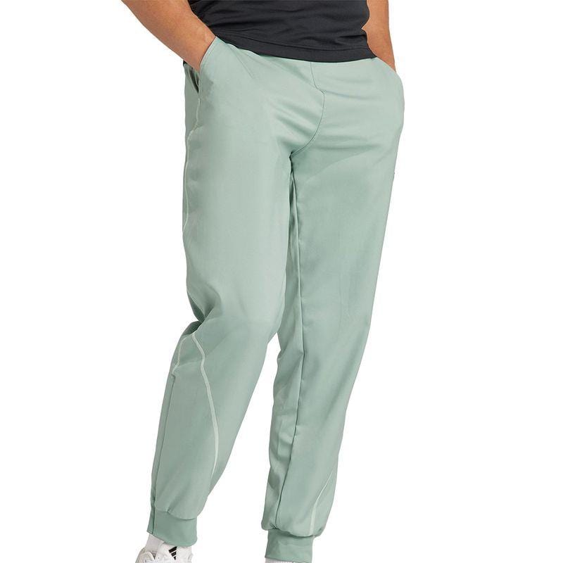 A person dressed in a pair of ADIDAS Melbourne Men's Pro Tennis Pants in green, with hands in pockets, combined with a black shirt and white sneakers. The pants feature white stitching near the ankles, adding both style and comfort. The scene is set against a plain white background.