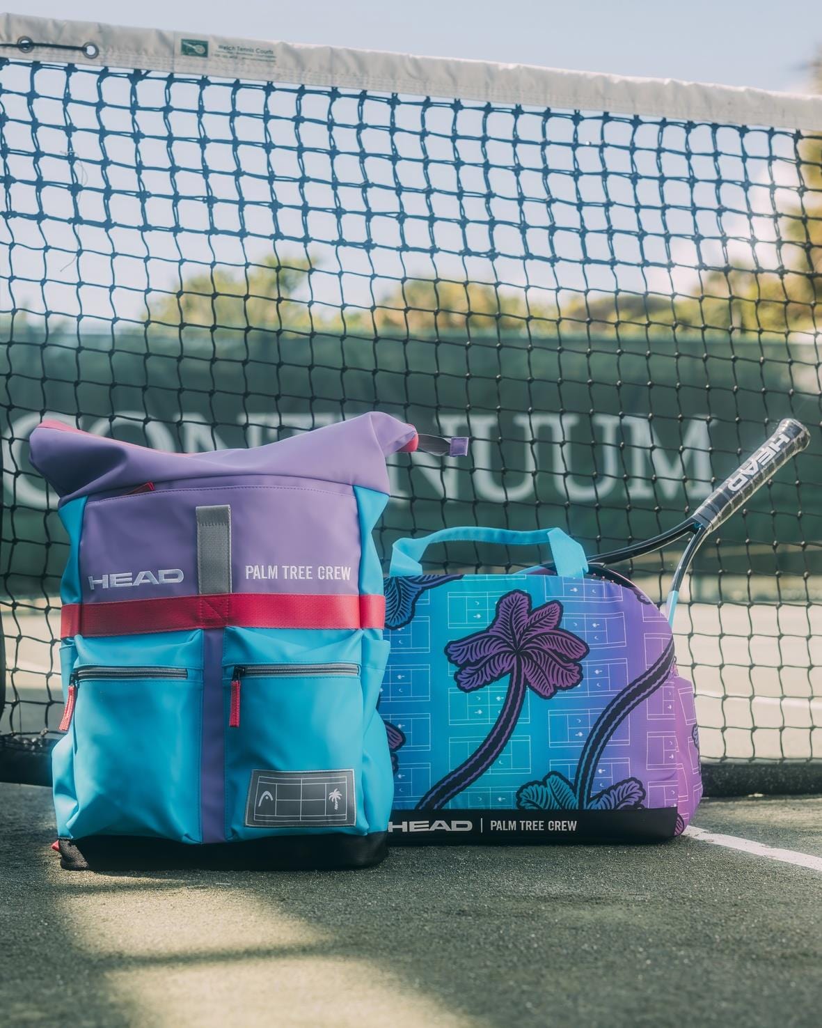 Two vibrant HEAD Tour Tennis Duffle Bags L - Palm Tree Crew are displayed on a tennis court in front of a net, with a tennis racket partly visible beside them.