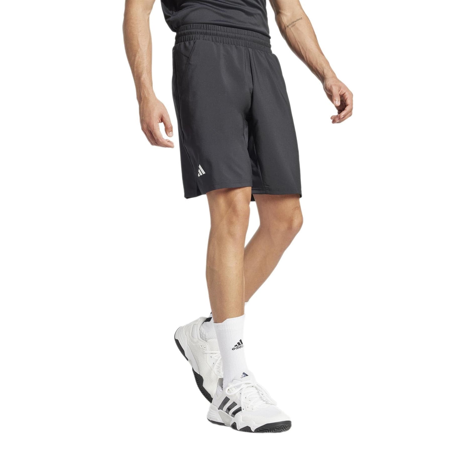 A person stands with slightly bent knees, wearing black adidas Men's 3 Stripe Tennis Shorts and a matching shirt, along with white Adidas Club sneakers featuring black stripes. Moisture-wicking socks complete the look, displaying logos on the shorts and socks to showcase only their lower body.