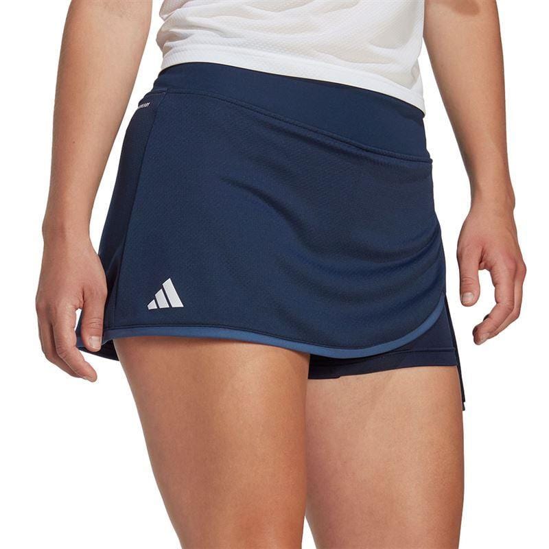 A person is wearing the ADIDAS Womens Club Tennis Skirt in navy, featuring the iconic adidas logo on the front. This tennis skirt boasts a wrap-style design and breathable AEROREADY fabric. The look is completed with a crisp white top, ensuring both style and performance on the court.