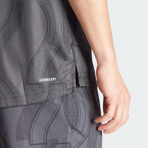 Close-up of a person wearing an adidas Men's Club Graphic Tennis T-Shirt in carbon black, showcasing textured patterns. The AEROREADY fabric label appears near the bottom, crafted from 100% recycled materials. The person's hand rests by their side against a plain light grey background.