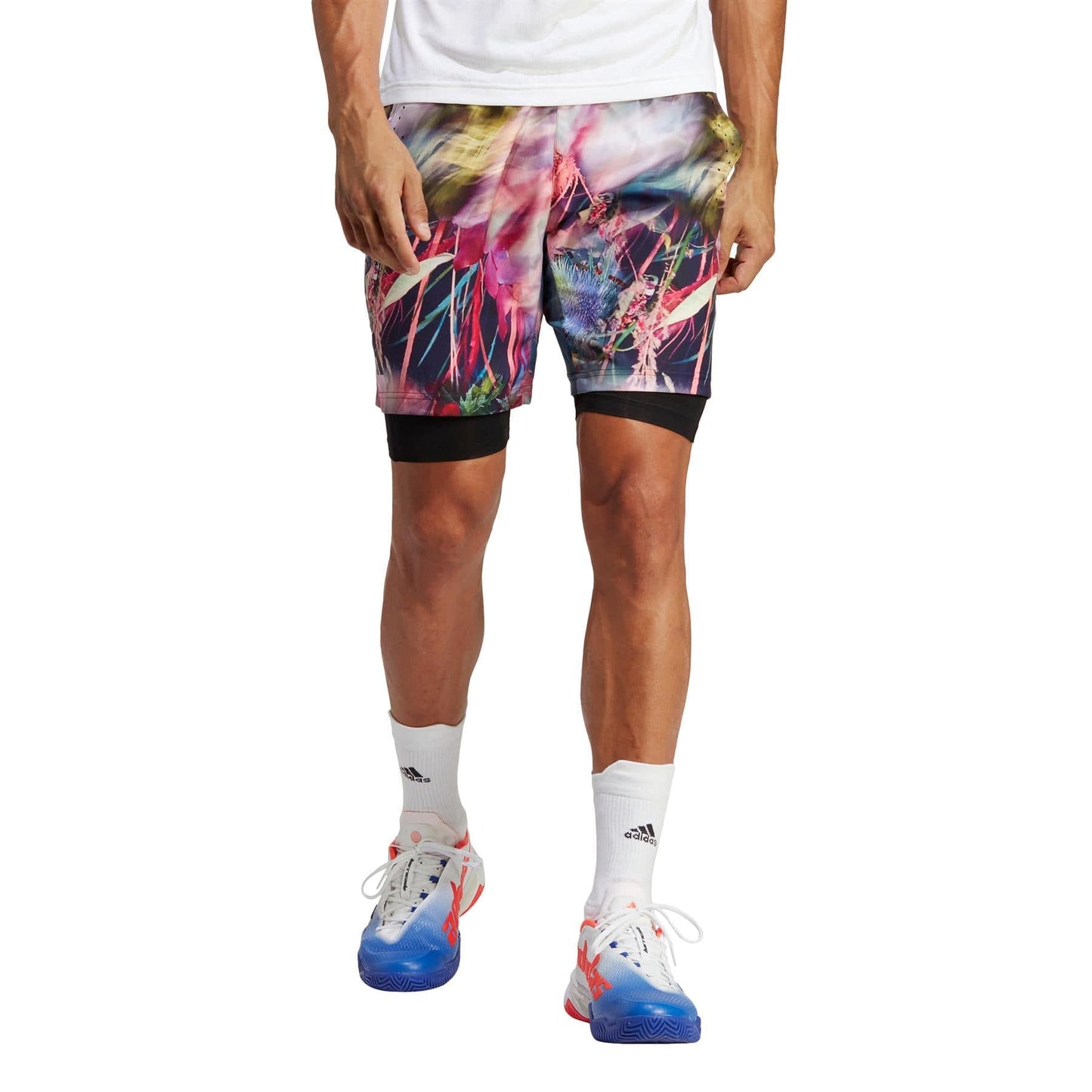 A person effortlessly flaunts a vibrant pair of adidas Melbourne Men's Ergo Graphic Tennis Shorts in Black and Multi colors, crafted from recycled materials, paired with white socks and blue athletic shoes. The shorts feature an abstract design in bright pink and blue hues against a plain background.