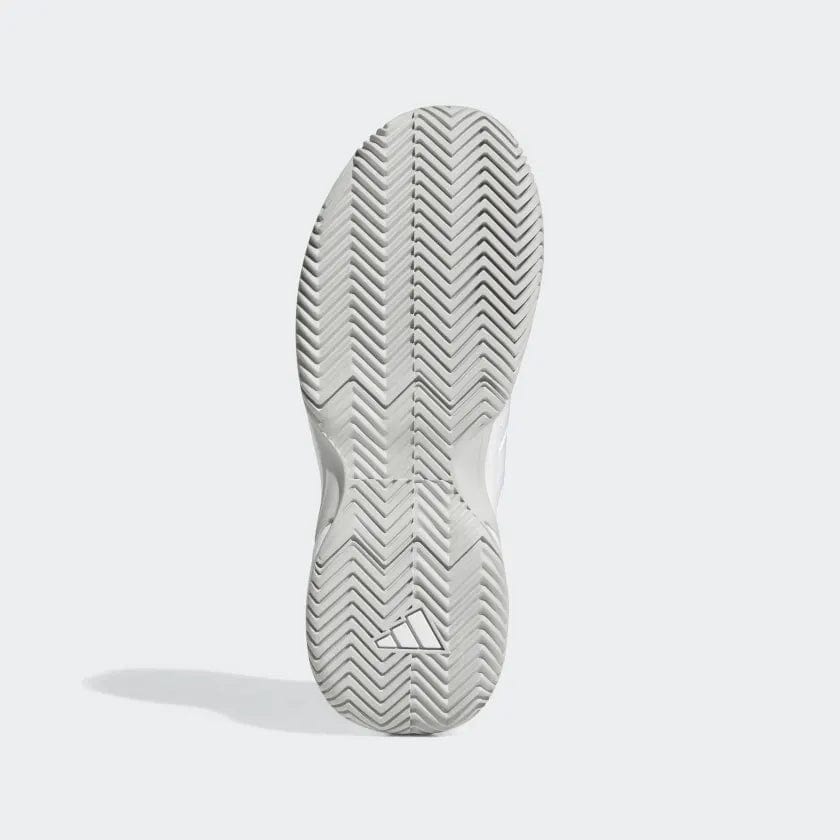 The image shows the sole of an adidas GameCourt 2 women's tennis shoe in white, featuring a herringbone tread pattern perfect for tennis play. It includes a subtle triangular adidas logo near the heel, with a shadow on the left suggesting it stands upright on a light surface, optimized for hard court outsoles.