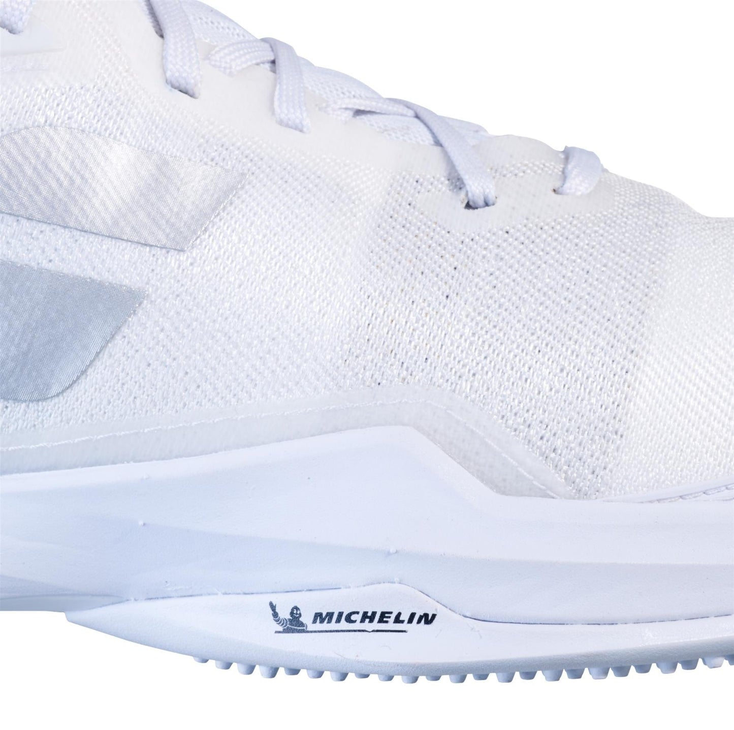 The Babolat Jet Mach 3 Grass Court Women's Wimbledon Tennis Shoes in White/Silver highlight a close-up view of a sleek athletic shoe with a textured surface and subtle geometric patterns. Featuring Matryx® technology for enhanced support, the design ensures durable traction with its Michelin sole, combining functionality with modern style.